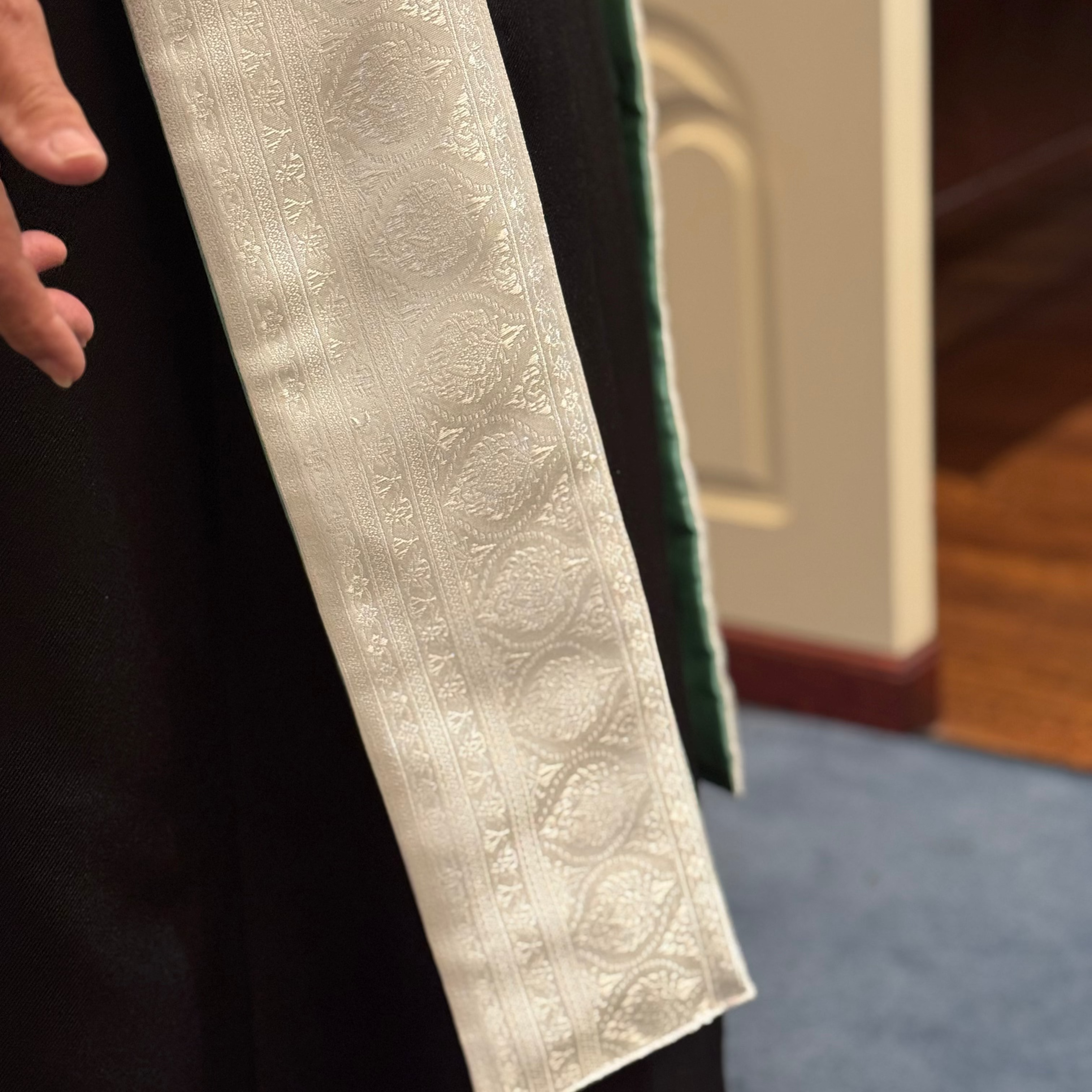 Person wearing a black robe with a white embroidered stole, blurred background