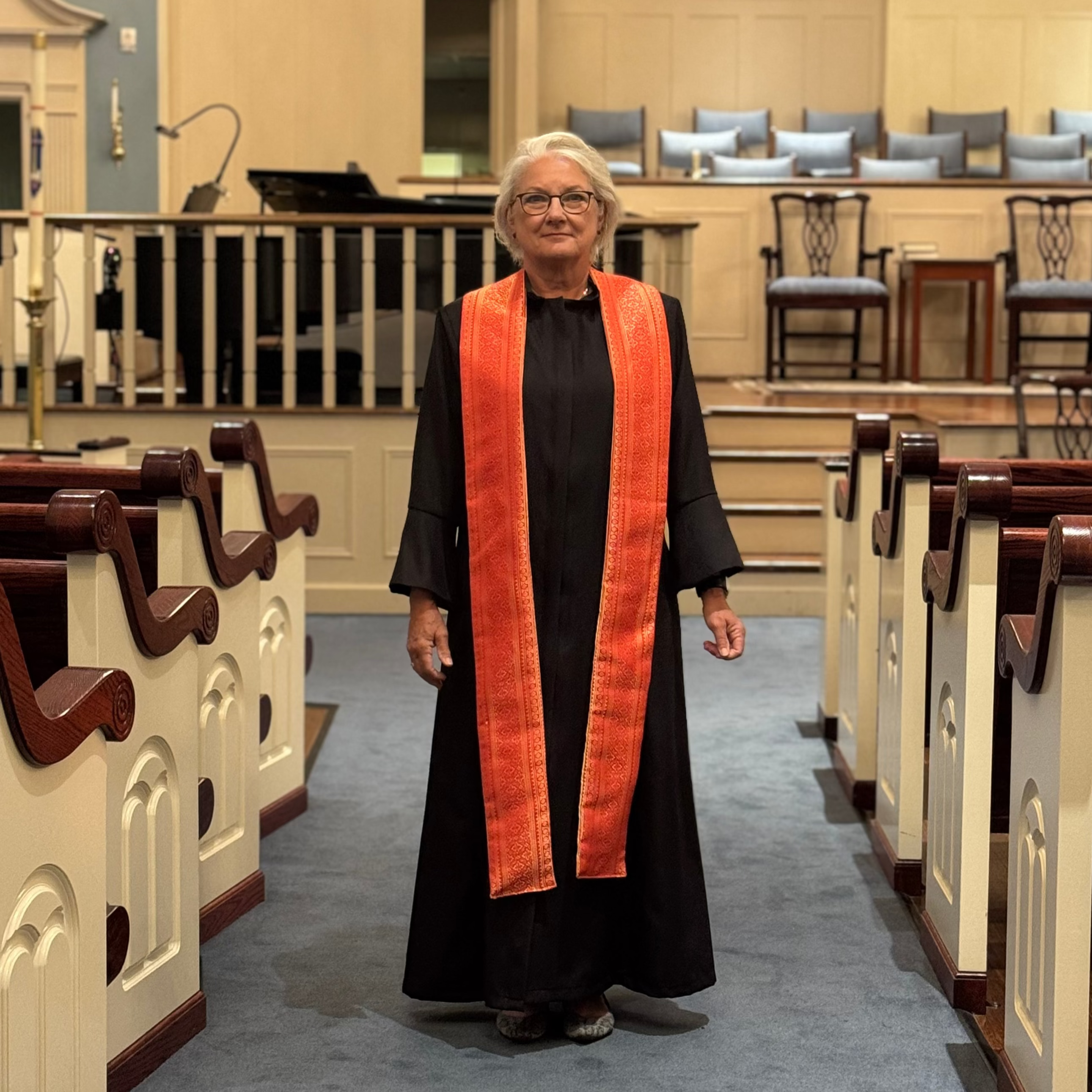 Person in a church wearing a long orange stole and black robes, standing in an aisle.
